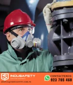Trabajador con respirador de media cara, casco rojo y lentes de seguridad realizando inspección industrial.